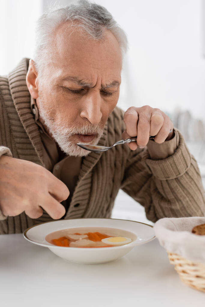 harmaa tukka mies Parkinsonin oireyhtymä syö keittoa päivälliseksi keittiössä - Valokuva, kuva