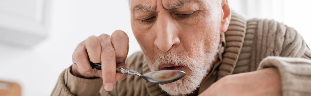 γενειοφόρος ηλικιωμένος άνδρας με παρκινσονικό σύνδρομο και τρέμουλο στα χέρια κρατώντας κουτάλι, ενώ έχοντας δείπνο στο σπίτι, πανό - Φωτογραφία, εικόνα