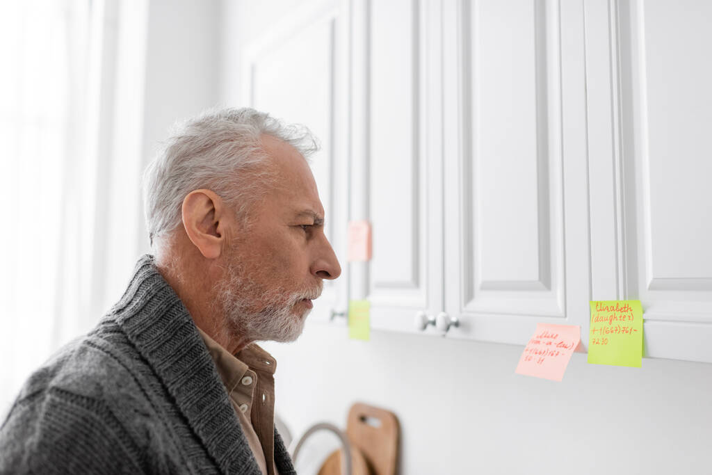 side view of grey haired man with alzheimer syndrome looking at sticky notes with names and phone numbers in kitchen - Photo, Image