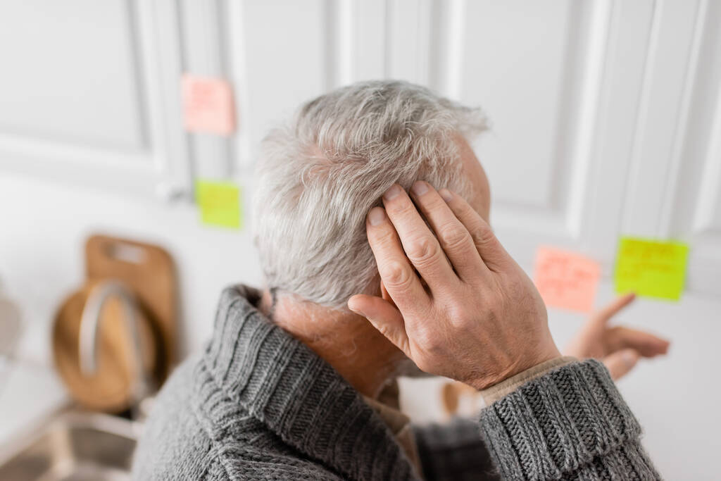 γκριζομάλλης ηλικιωμένος άνδρας με σύνδρομο Αλτσχάιμερ που αγγίζει το κεφάλι και δείχνει με θολή αυτοκόλλητη σημείωση στην κουζίνα - Φωτογραφία, εικόνα