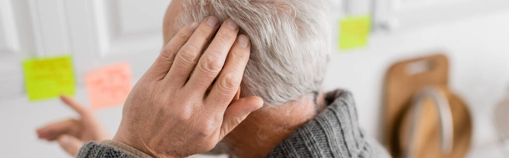 aged man with alzheimer disease touching head and pointing at blurred sticky notes in kitchen, banner - Foto, afbeelding