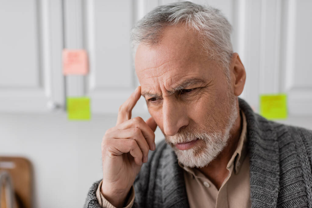 thoughtful senior man with alzheimer disease touching head near blurred sticky notes in kitchen - Photo, Image