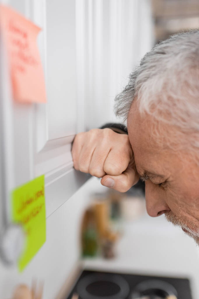 side view of depressed senior man suffering from memory loss while standing near blurred sticky notes in kitchen - Foto, Imagem