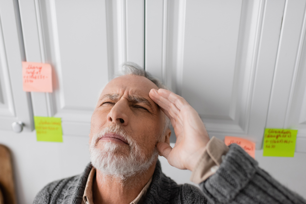 senior man with closed eyes and hand near head standing near blurred sticky notes while suffering from memory loss - Foto, afbeelding
