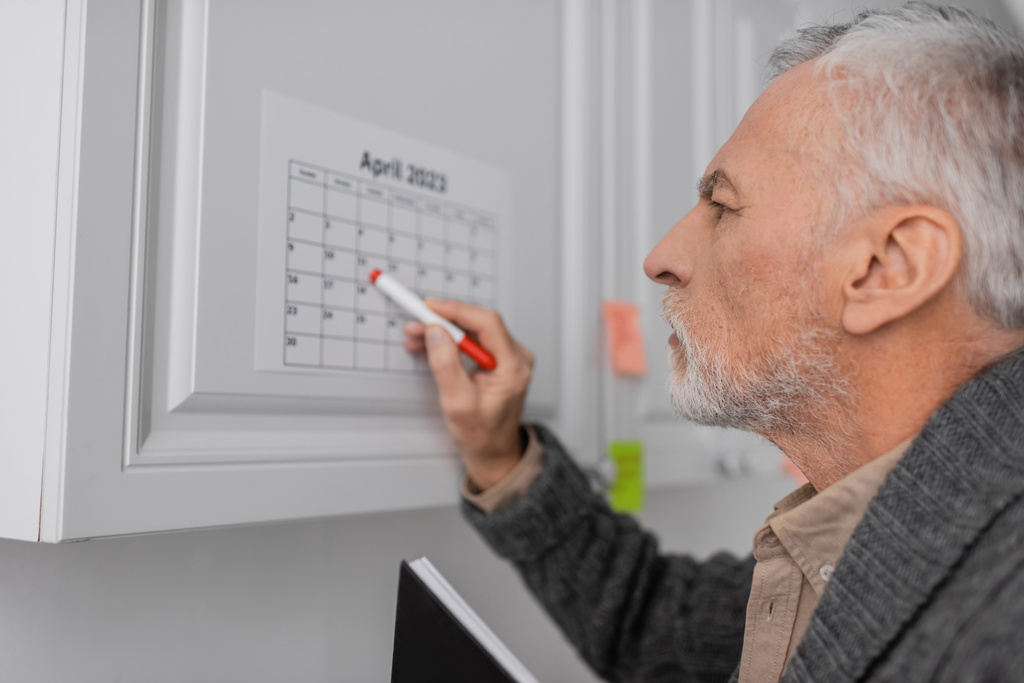 Вид сбоку пожилого человека с альфа-синдромом, указывающего войлочной ручкой на календарь на кухне - Фото, изображение