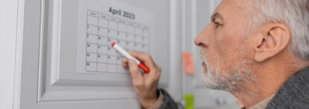 side view of senior man with alzheimer disease pointing with felt pen at calendar in kitchen, banner - Fotó, kép