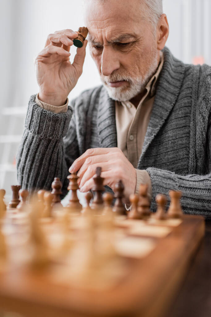 aged man sick on alzheimer syndrome holding figure while thinking near chessboard on blurred foreground - Fotó, kép