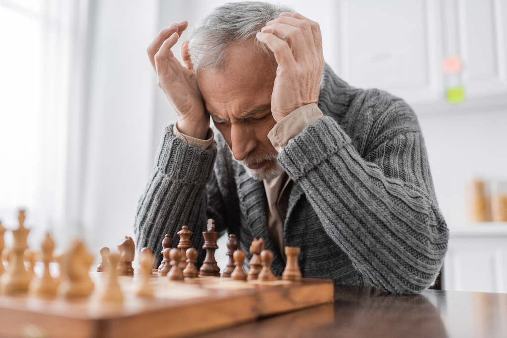 stressed man suffering from alzheimer syndrome and sitting at chessboard with closed eyes and hands near head  - Foto, immagini