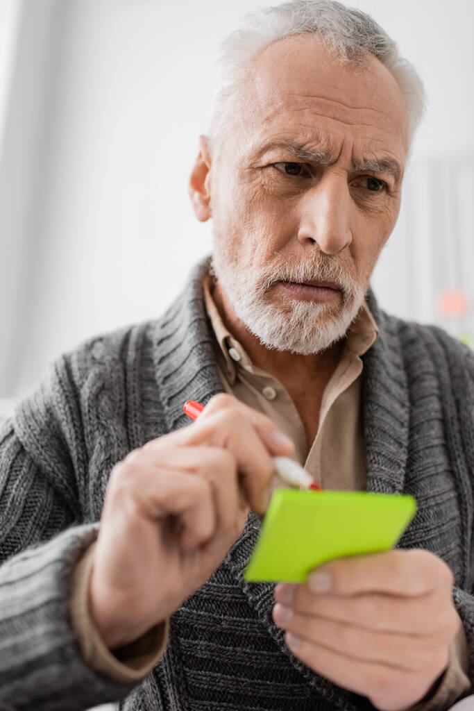 pensive senior man holding felt pen and sticky notes while suffering from memory loss  - Foto, afbeelding