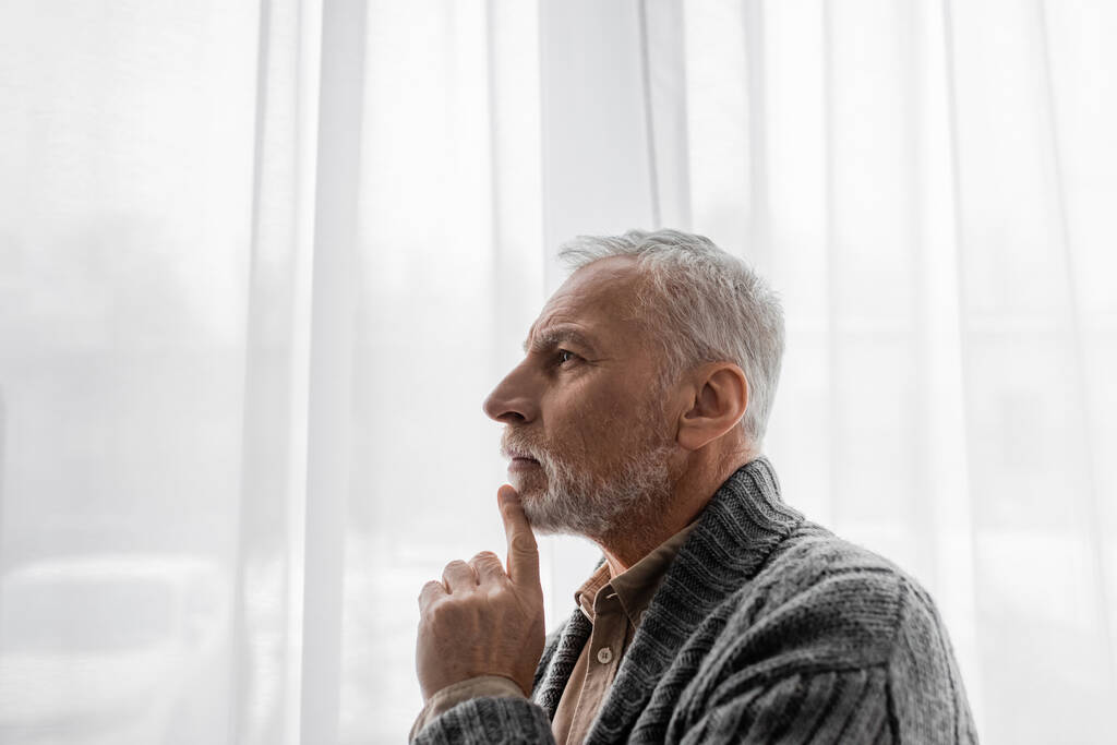 πλαϊνή άποψη του ηλικιωμένου άνδρα με νόσο Αλτσχάιμερ κοιτάζοντας μακριά κοντά στο παράθυρο στο σπίτι - Φωτογραφία, εικόνα