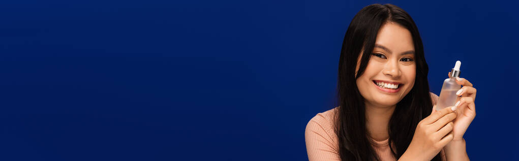 Улыбающаяся брюнетка азиатка, держащая косметическую сыворотку изолированную на голубом, баннер  - Фото, изображение