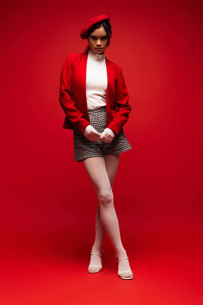 Full length of trendy asian model in tights and gloves standing on red background  - Foto, Imagem