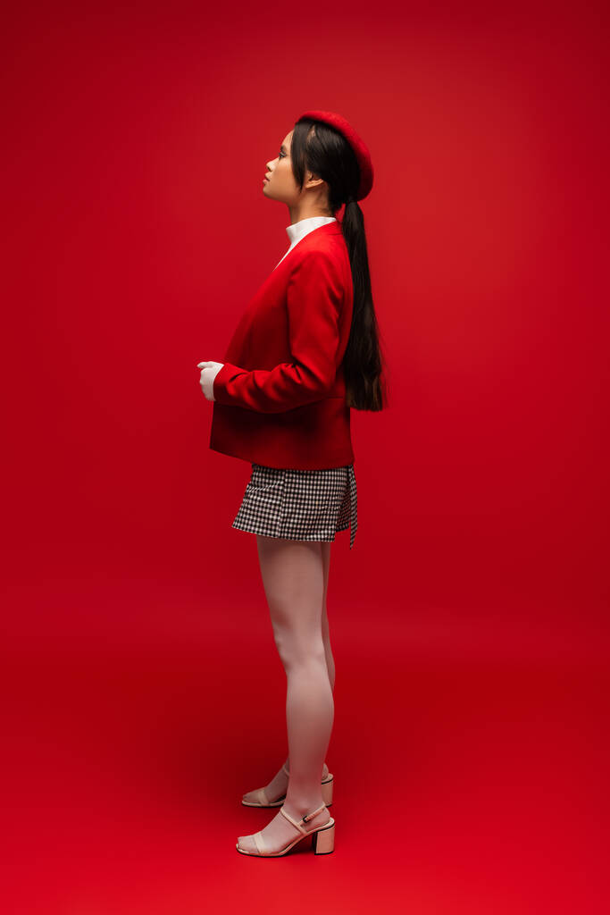 サイドビューのスタイリッシュなアジアの女性で白い手袋とタイツは赤の背景に立って  - 写真・画像