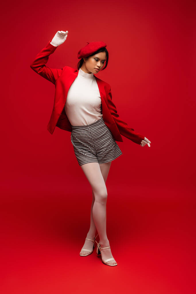 Full length of trendy asian woman in beret and blazer posing on red background  - Foto, Imagen