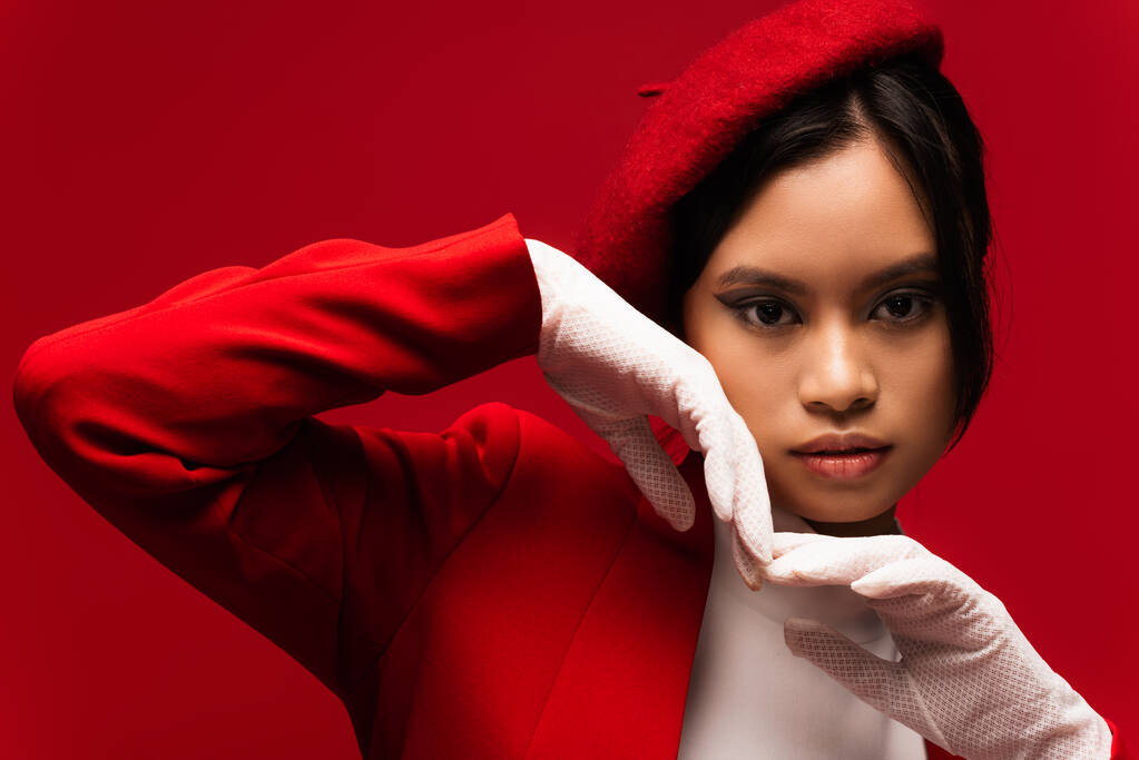 Portrait of asian model in beret and gloves holding hands near face isolated on red  - Foto, imagen