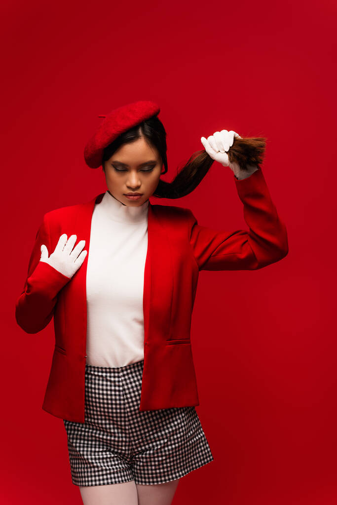 Stylish asian model in beret touching hair while standing isolated on red  - Photo, image