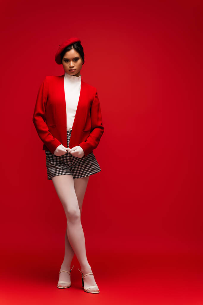 Full length of stylish asian woman in jacket and tights looking at camera on red background  - Photo, image