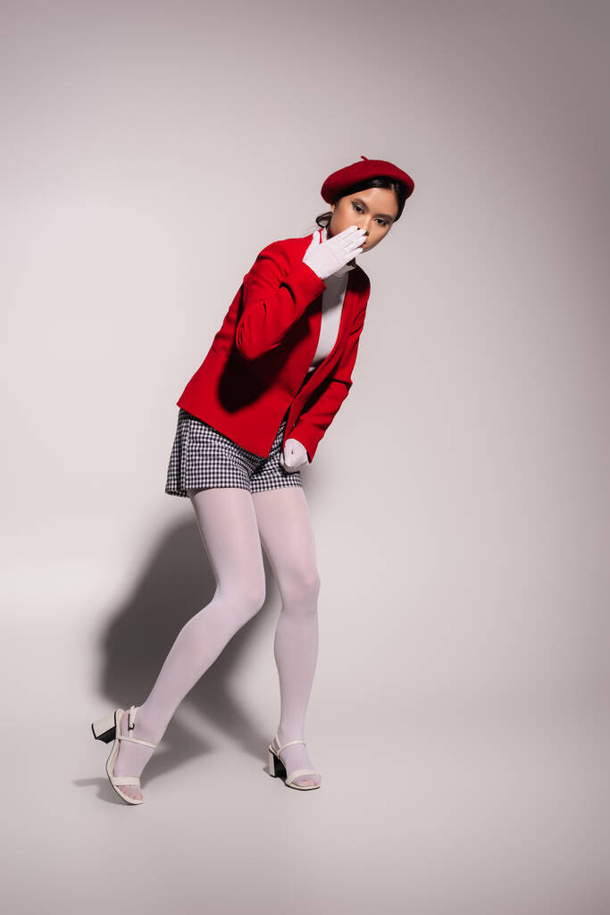 Stylish asian woman in red beret and gloves holding hand near mouth on grey background  - Foto, Imagem