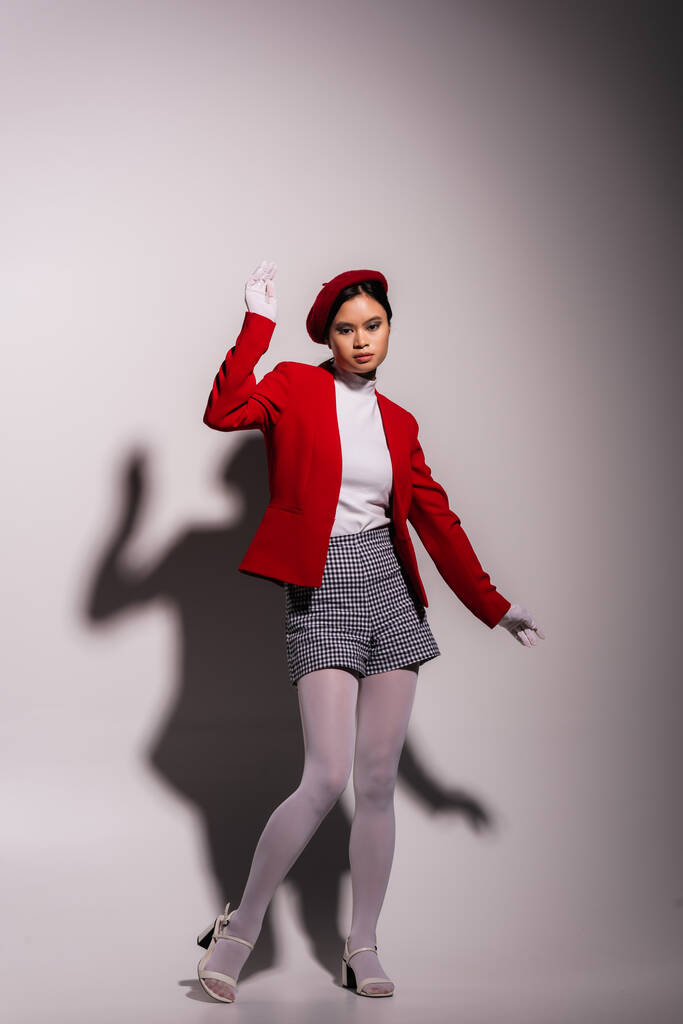 Full length of stylish asian model in red beret and jacket standing on grey background with shadow  - Foto, imagen