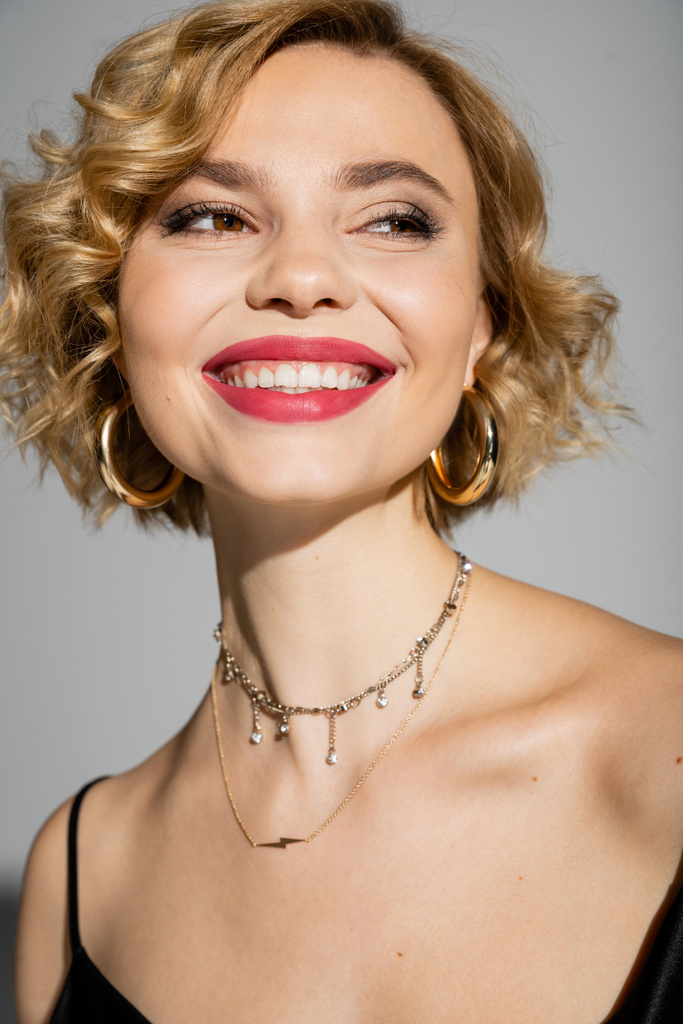 portrait of happy young woman in hoop earrings and silver necklace smiling isolated on grey  - Fotografie, Obrázek