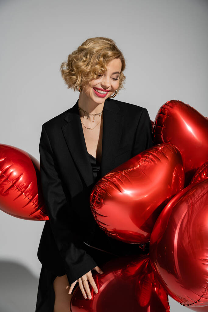 positive blonde woman in black blazer and slip dress standing with red balloons isolated on grey  - Foto, Imagen