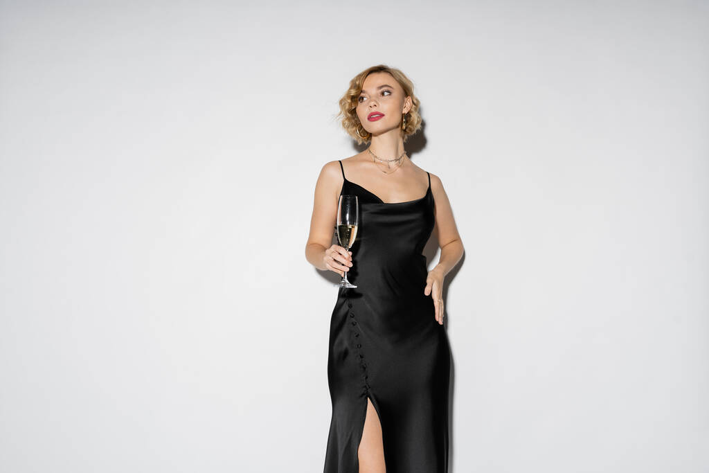 blonde woman in satin slip dress holding glass of champagne while standing with hand on hip isolated on grey  - Fotografie, Obrázek