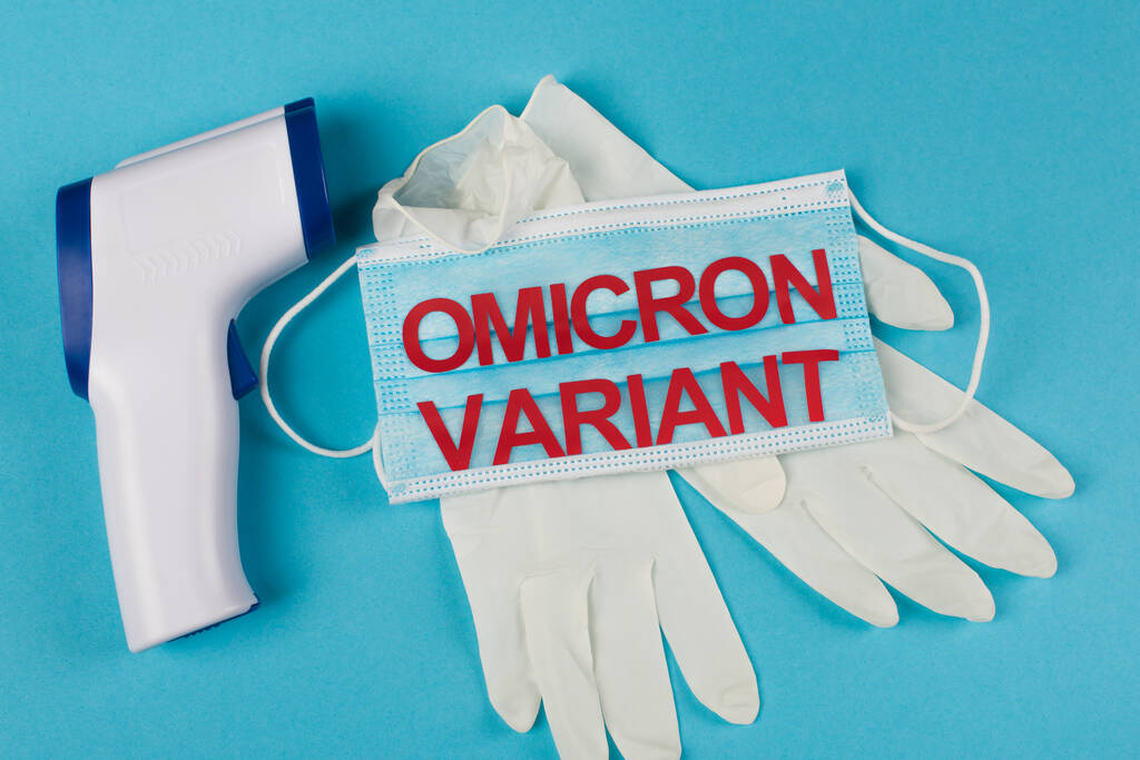Top view of medical mask with omicron variant lettering near latex gloves and pyrometer on blue background  - Φωτογραφία, εικόνα