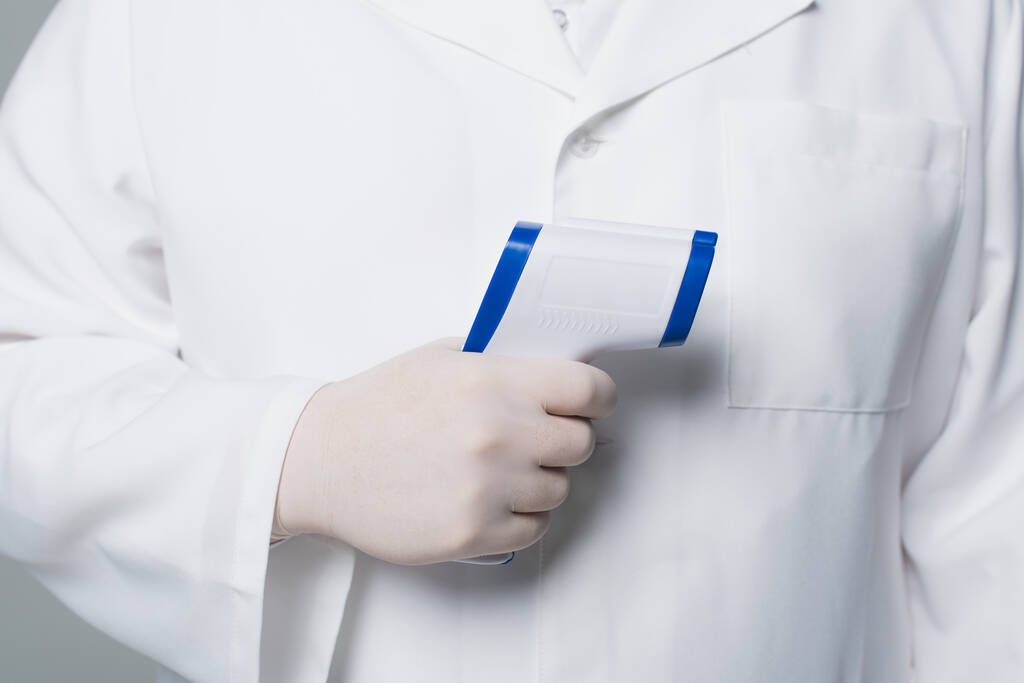 Обрезанный вид врача в белом халате и латексной перчатке с пирометром, изолированным на сером  - Фото, изображение
