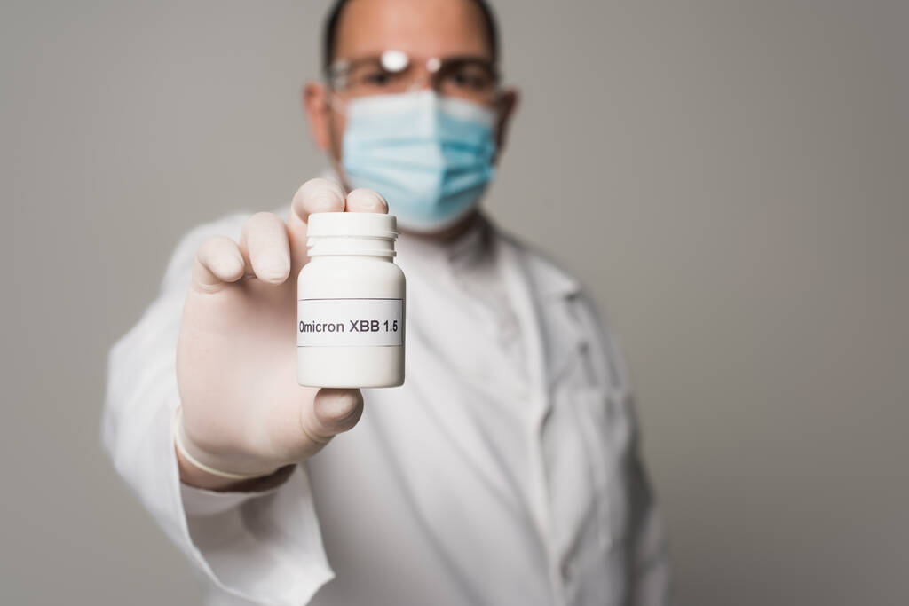 Размытый ученый в медицинской маске держит таблетки с омикронными надписями изолированы на сером  - Фото, изображение