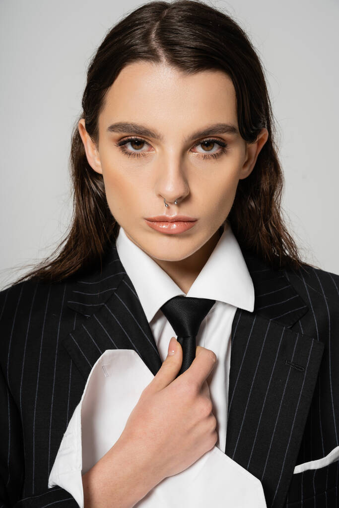 portrait of stylish brunette woman in black blazer and white shirt touching tie and looking at camera isolated on grey - Φωτογραφία, εικόνα