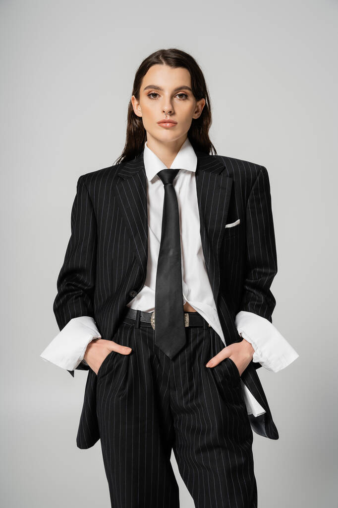front view of brunette woman in stylish oversize suit holding hands in pockets while looking at camera isolated on grey - Photo, Image