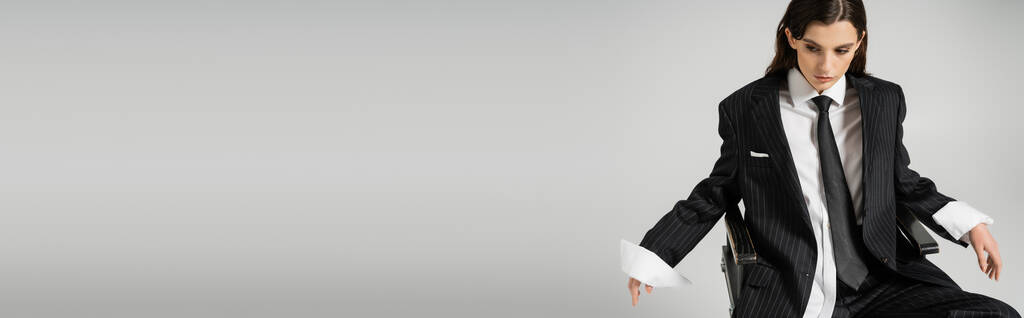 Nuori nainen valkoisessa paidassa ja musta bleiseri solmio katselee alas istuessaan eristetty harmaa, banneri - Valokuva, kuva