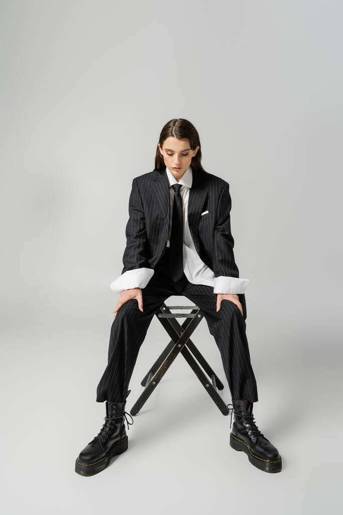 full length of pretty model in black elegant suit and rough leather boots sitting on chair on grey background - Foto, imagen