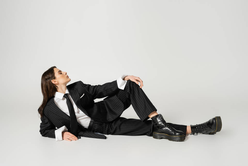 full length of young woman in stylish formal wear and laced-up boots lying on grey background - Fotoğraf, Görsel
