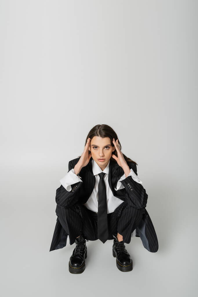 brunette woman in black striped suit and rough boots holding hands near face and looking at camera while sitting on grey  - Foto, immagini
