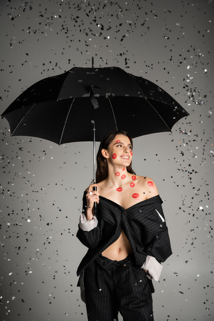 sexy and stylish woman standing with hand in pocket under black umbrella and shiny confetti on grey background - Photo, Image