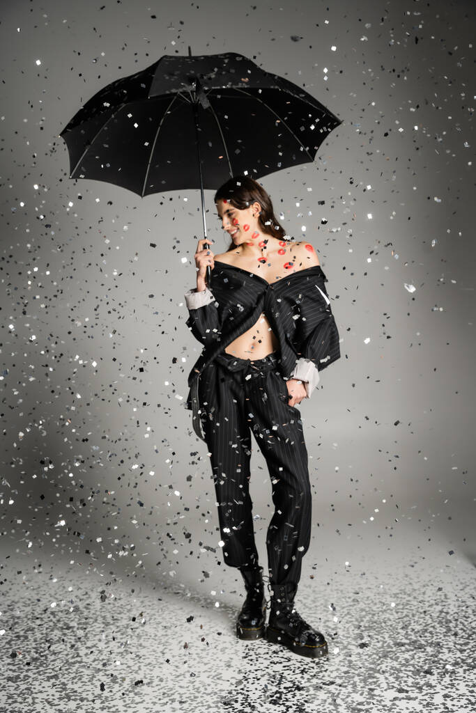 happy woman in black stylish clothes standing with umbrella under sparkling confetti on grey  - Foto, imagen