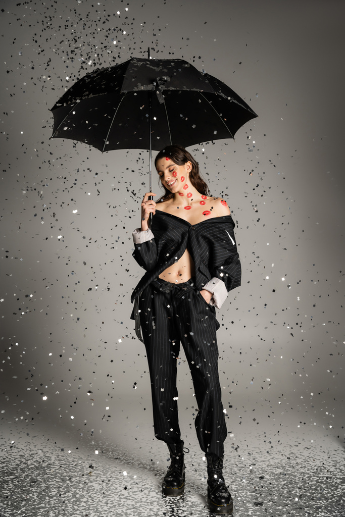 happy and stylish woman with red lip prints on body standing with black umbrella under shiny confetti on grey background - Foto, afbeelding