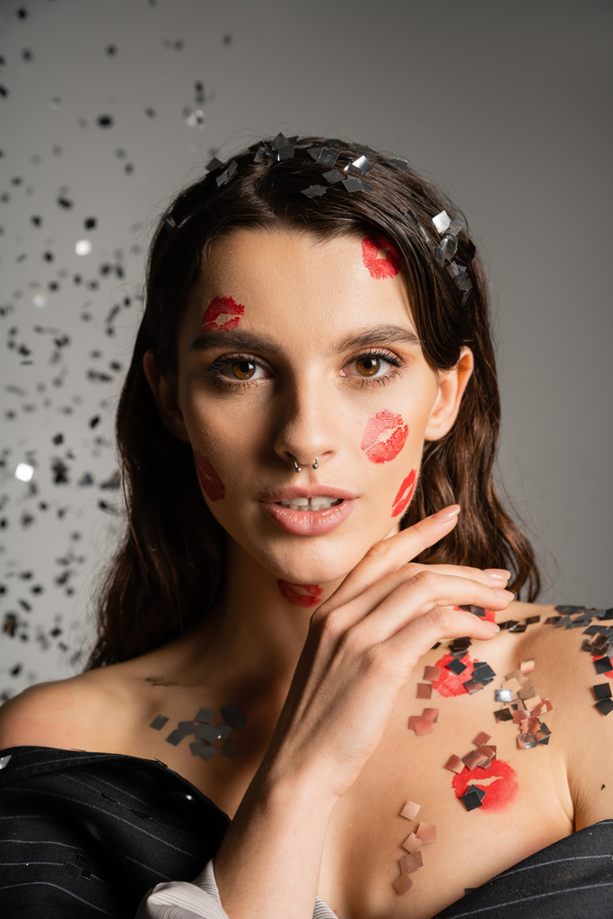 charming woman with red kiss prints on face looking at camera near sparkling confetti on grey  - Photo, image