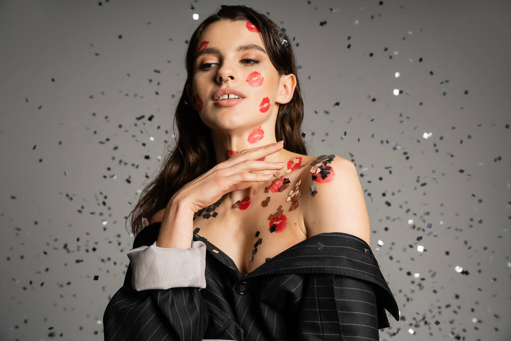brunette woman in black blazer with bare shoulders and lip prints on body standing under shiny confetti on grey background - Photo, image