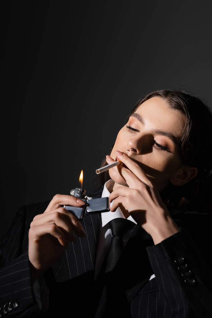 молода і елегантна жінка в формальному носінні освітлення сигарети ізольовані на чорному
 - Фото, зображення