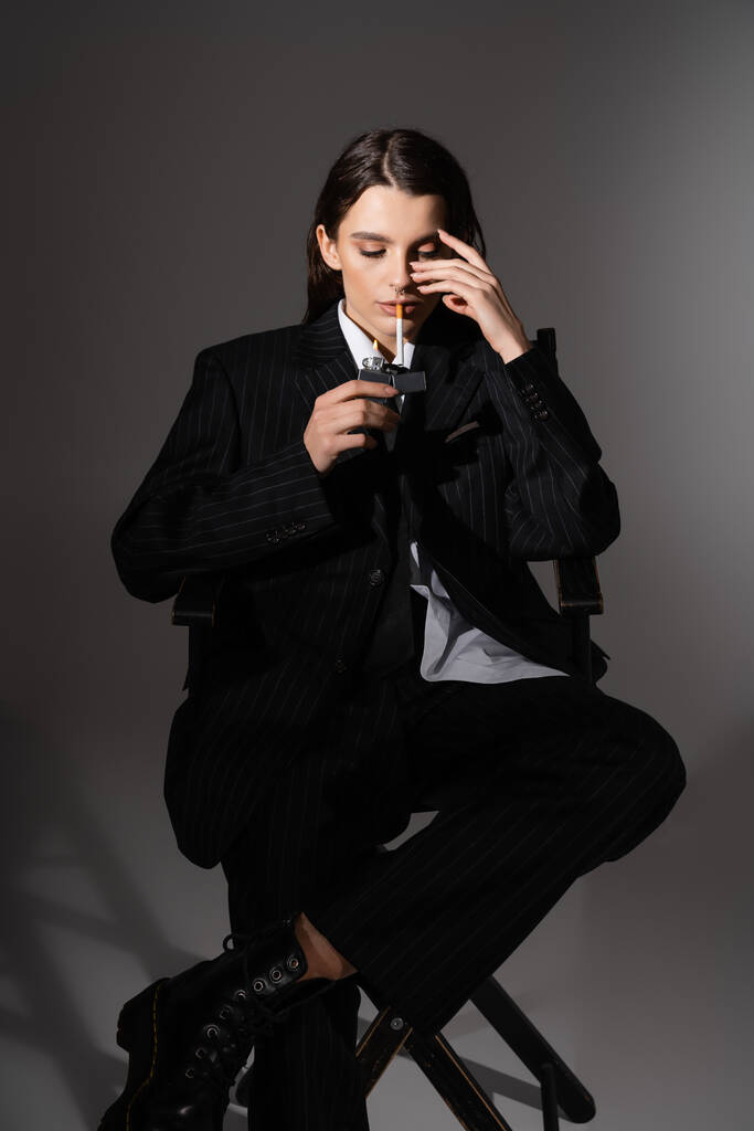fashionable woman in black pantsuit lighting cigarette while sitting on dark grey  - Foto, imagen