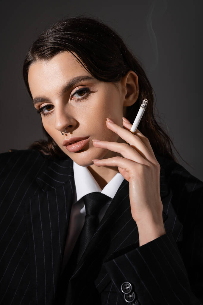 портрет брюнетки в чорному смугастому блістері, що тримає сигарету і дивиться на камеру ізольовано на темно-сірому
 - Фото, зображення
