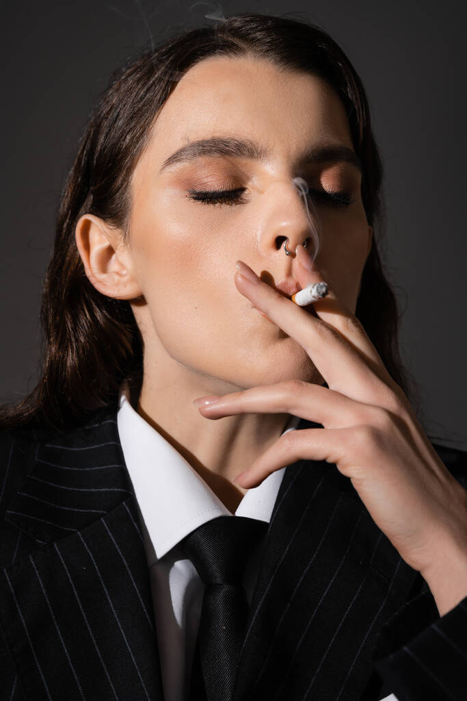 portrait of young woman in stylish formal wear smoking with closed eyes isolated on dark grey - Foto, imagen