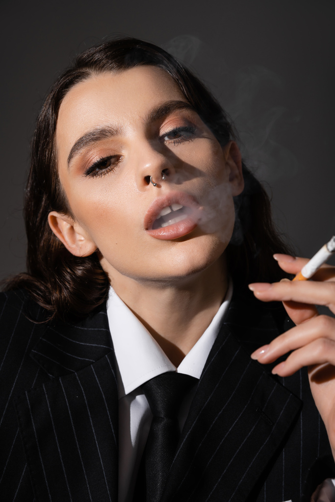 portrait of elegant woman with makeup and piercing looking at camera while smoking isolated on dark grey - Foto, afbeelding