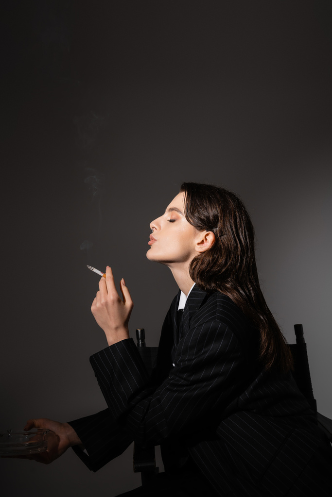 side view of brunette woman in black blazer holding ashtray and smoking on dark grey background - Photo, image