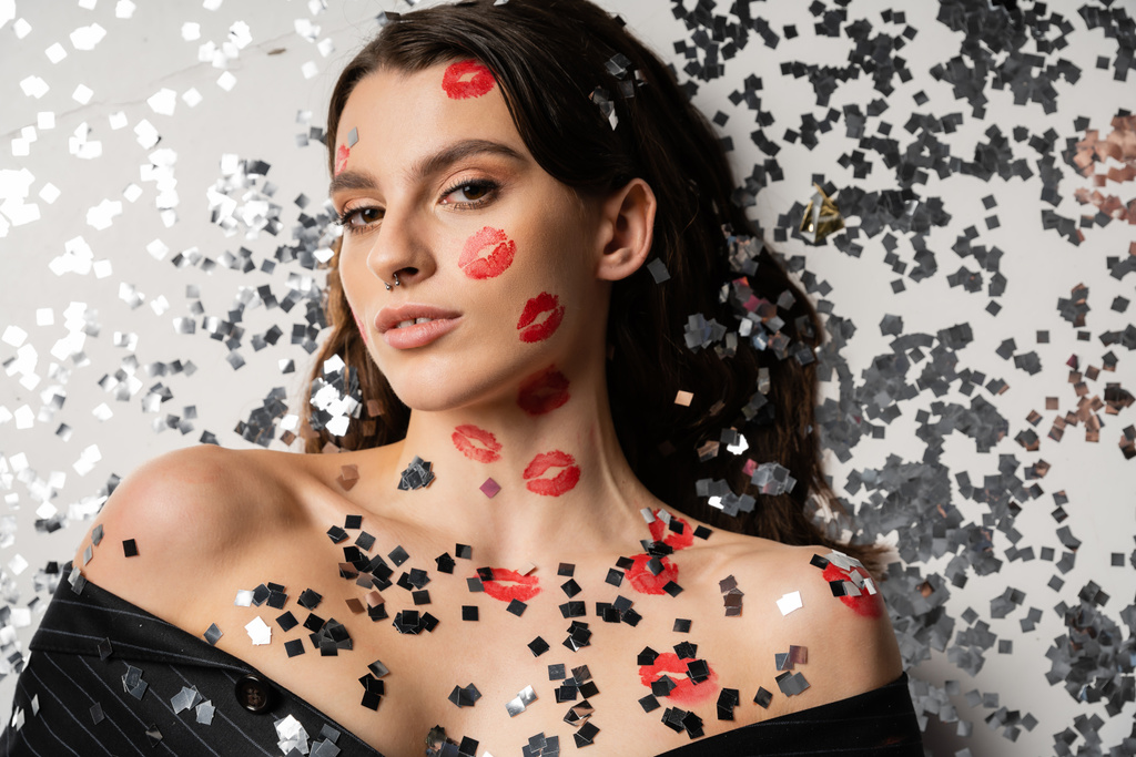 top view of brunette woman with red kiss prints looking at camera while lying near shiny confetti on grey background - Photo, Image