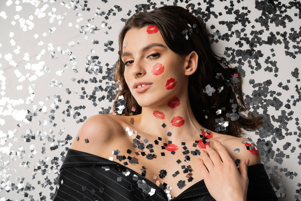 top view of stylish woman with red lip prints lying near festive confetti and looking at camera on grey background - Photo, image