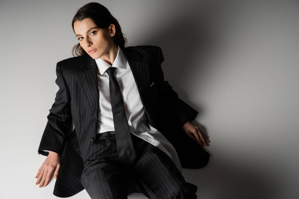 high angle view of stylish model in white shirt and black oversize suit looking at camera while sitting on grey background - Φωτογραφία, εικόνα