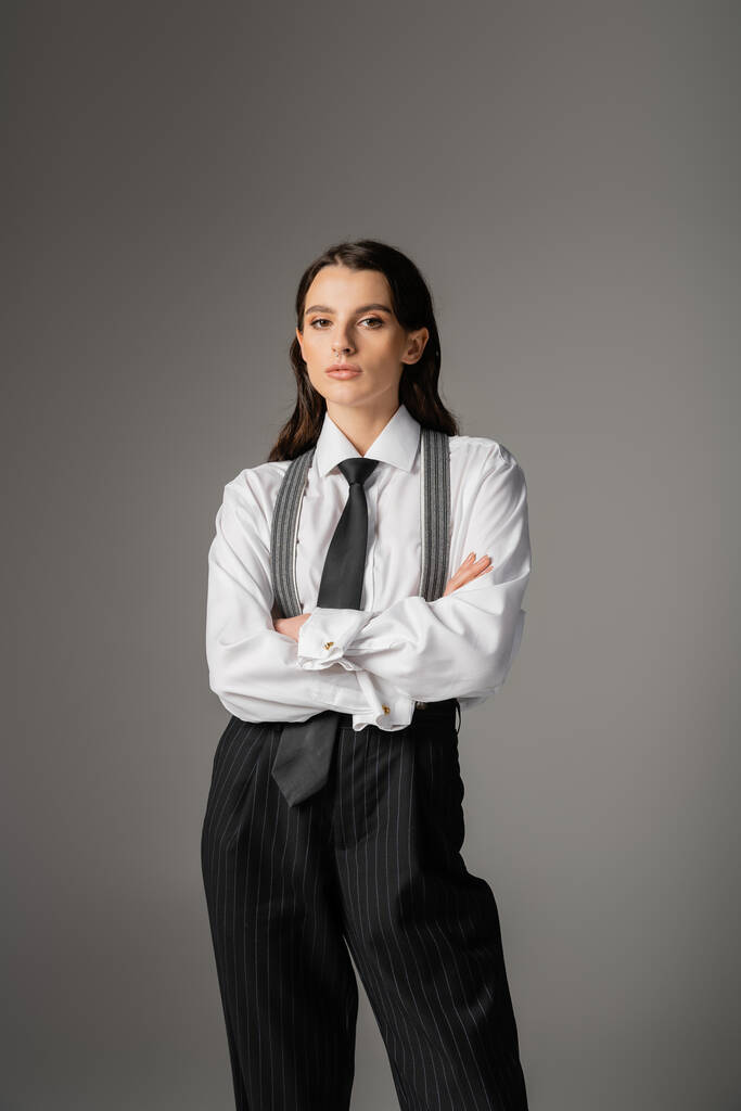 stylish woman in white shirt and trousers with suspenders standing with crossed arms isolated on grey - Photo, image